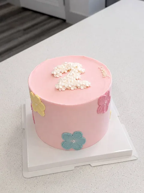 Pink single-tier cake with a white flower cluster and pastel flower accents.